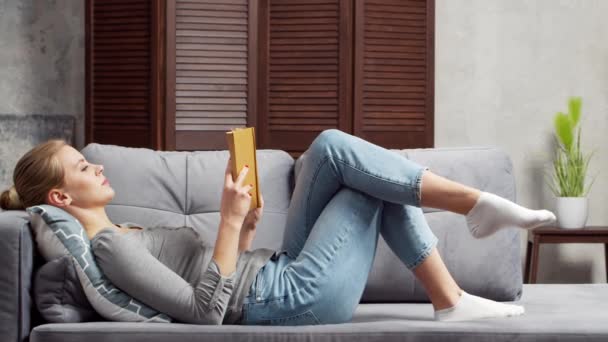 금발 소녀는 책이나 책을 읽고 있습니다. 아름다운 여인 이 거실에서 쉬고 쉬고 있다. — 비디오