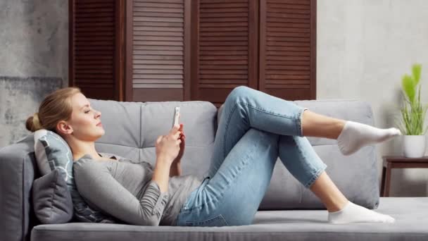 Mujer joven trabaja con un teléfono inteligente acostado en el sofá. Estudiante, empresaria o freelancer que trabaja o estudia de forma remota a través de Internet. Enseñanza a distancia y concepto de trabajo. — Vídeos de Stock