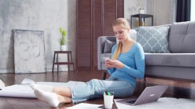 Kadın, evde yerde otururken akıllı telefon ve dizüstü bilgisayar kullanarak belgelerle çalışıyor. Öğrenci, girişimci ya da serbest çalışan ya da uzaktan eğitim gören bir kız. Mesafe öğrenme ve iş kavramı.