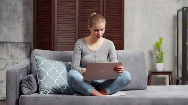 Jovem trabalha usando um laptop enquanto se senta no sofá. Estudante, empreendedora ou freelancer trabalhando ou estudando remotamente via internet. Ensino à distância e conceito de emprego. — Vídeo de Stock