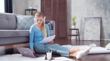 Genç bir kadın evde yerde otururken dizüstü bilgisayar kullanarak belgelerle çalışıyor. Öğrenci, girişimci ya da serbest çalışan ya da uzaktan eğitim gören bir kız. Mesafe öğrenme ve iş kavramı.