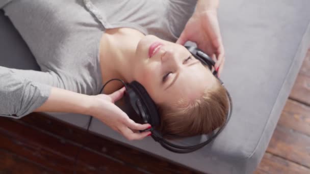 Jovem menina bonita encontra-se em um sofá em fones de ouvido e ouve música. Mulher loira está descansando em casa. — Vídeo de Stock