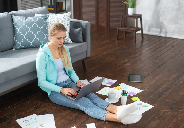 Junge Frau arbeitet mit Laptop mit Dokumenten, während sie zu Hause auf dem Boden sitzt. Studentin, Unternehmerin oder Freiberuflerin, die aus der Ferne über das Internet arbeitet oder studiert. — Stockfoto