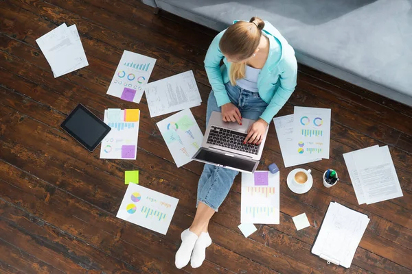 Jovem trabalha com documentos usando um laptop enquanto está sentado no chão em casa. Estudante, empreendedora ou freelancer trabalhando ou estudando remotamente via internet. — Fotografia de Stock