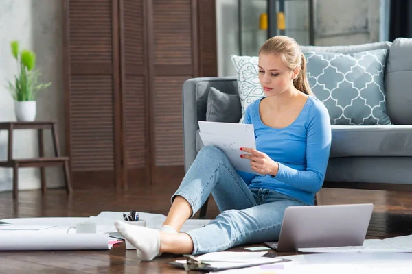 Mujer joven trabaja con documentos utilizando un ordenador portátil mientras está sentado en el suelo en casa. Estudiante, empresaria o freelancer que trabaja o estudia de forma remota a través de Internet. — Foto de Stock