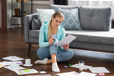 Genç bir kadın evde yerde otururken belgelerle çalışıyor. Öğrenci, girişimci veya serbest çalışan kız İnternet üzerinden çalışıyor veya uzaktan öğrenim görüyor.