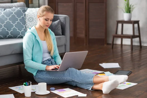 年轻妇女坐在家里的地板上，用笔记本电脑处理文件。通过因特网从事远程工作或学习的学生、企业家或自由职业者. — 图库照片