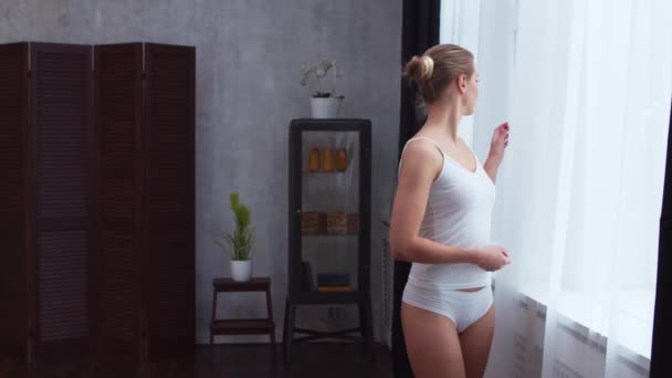 Ung kvinna i vita underkläder står framför fönstret och tittar in i det. Flicka hemma i vardagsrummet. — Stockvideo