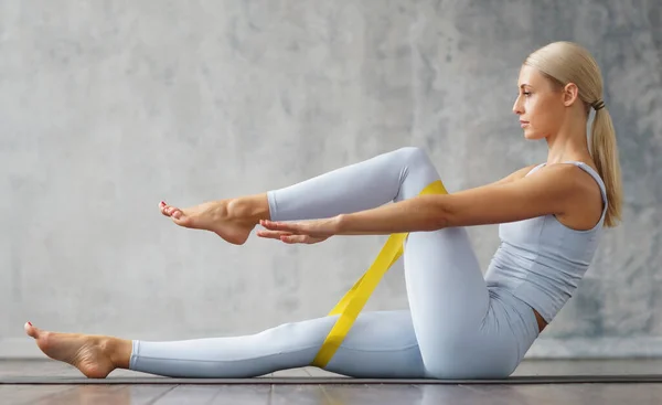 Young slender blonde woman in sportswear is engaged in fitness at home. Girl is using resistance band. Sport, health and lifestyle concept. — Fotografia de Stock