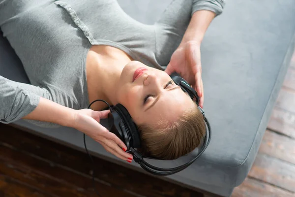 Das junge schöne Mädchen liegt mit Kopfhörern auf einem Sofa und hört Musik. Glückliche blonde Frau ruht sich zu Hause aus. — Stockfoto