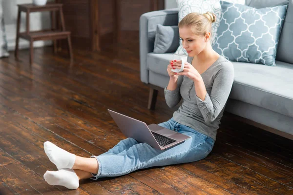 Молодая женщина работает с ноутбуком, сидя дома на полу. Студентка, предприниматель или девушка-фрилансер, работающая или обучающаяся дистанционно через интернет. — стоковое фото