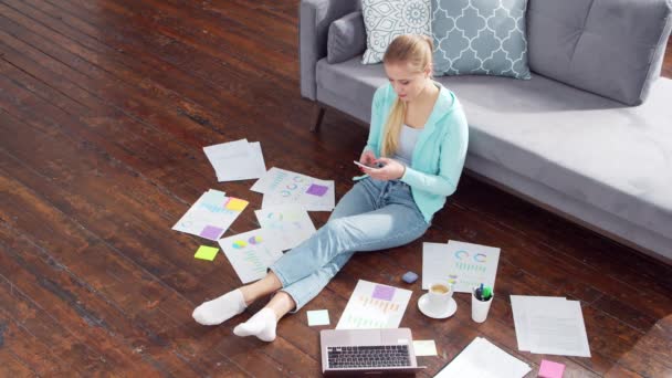 La mujer trabaja con documentos usando un teléfono inteligente y un portátil mientras está sentada en el suelo en casa. Estudiante, empresaria o freelancer que trabaja o estudia a distancia. Enseñanza a distancia y concepto de trabajo. — Vídeos de Stock