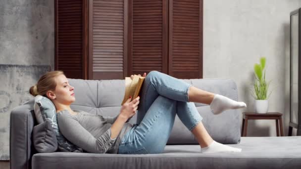 Chica rubia joven está leyendo un libro o un libro de texto. Una hermosa mujer está descansando y relajándose en casa en la sala de estar. — Vídeo de stock