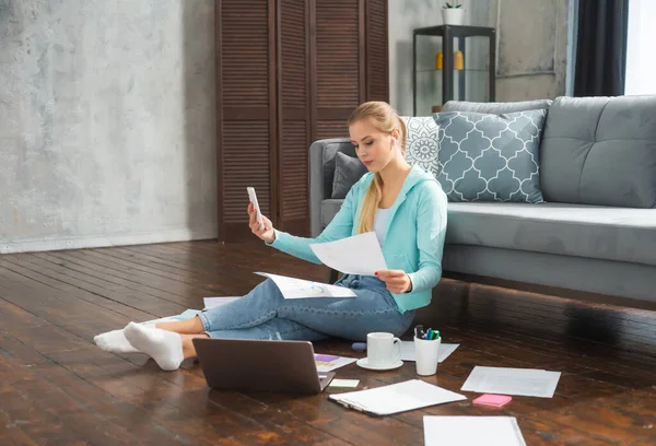 Молодая женщина работает с документами, используя смартфон и ноутбук, сидя дома на полу. Студентка, предприниматель или девушка-фрилансер, работающая или обучающаяся дистанционно через интернет. — стоковое фото
