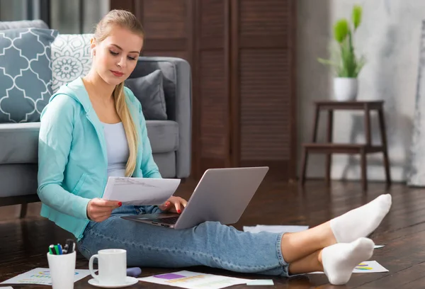 Junge Frau arbeitet mit Laptop mit Dokumenten, während sie zu Hause auf dem Boden sitzt. Studentin, Unternehmerin oder Freiberuflerin, die aus der Ferne über das Internet arbeitet oder studiert. — Stockfoto