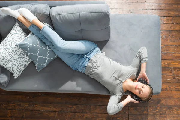 Das junge schöne Mädchen liegt mit Kopfhörern auf einem Sofa und hört Musik. Glückliche blonde Frau ruht sich zu Hause aus. — Stockfoto