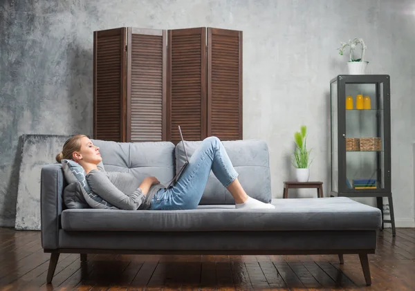 Die junge Frau arbeitet mit einem Laptop, der auf der Couch liegt. Studentin, Unternehmerin oder Freiberuflerin, die aus der Ferne über das Internet arbeitet oder studiert. — Stockfoto