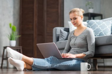 Genç bir kadın evde yerde otururken dizüstü bilgisayar kullanıyor. Öğrenci, girişimci veya serbest çalışan kız İnternet üzerinden çalışıyor veya uzaktan öğrenim görüyor.