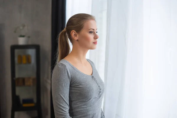 Young teenage girl is standing in front of the window and looking into it. Blond woman at home. — Stock Photo, Image