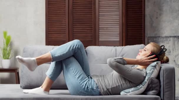 Joven chica hermosa se encuentra en un sofá en los auriculares y escucha música. Mujer feliz está descansando en casa. — Vídeos de Stock