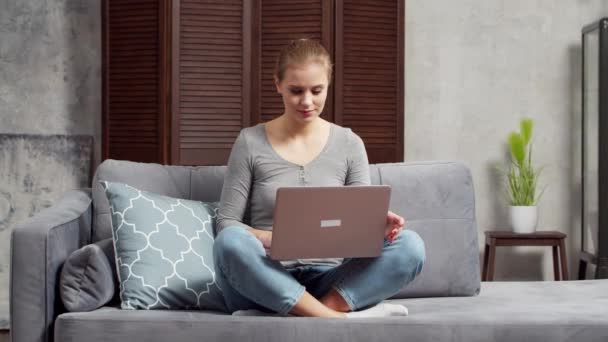 Mujer joven trabaja usando un ordenador portátil mientras está sentado en el sofá. Estudiante, empresaria o freelancer que trabaja o estudia de forma remota a través de Internet. Enseñanza a distancia y concepto de trabajo. — Vídeos de Stock