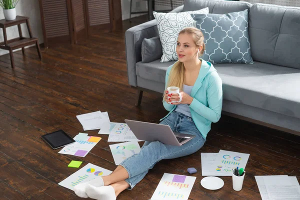 Молода жінка працює з документами, використовуючи ноутбук, сидячи на підлозі вдома. Студент, підприємець або дівчина-фрілансер, яка працює або навчається дистанційно через Інтернет . — стокове фото