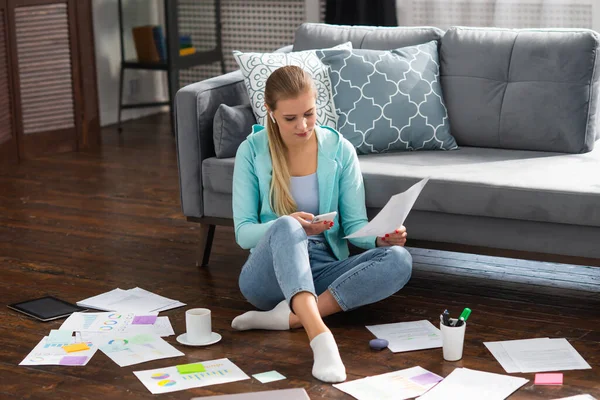 Junge Frau arbeitet mit Dokumenten per Smartphone, während sie zu Hause auf dem Boden sitzt. Studentin, Unternehmerin oder Freiberuflerin, die aus der Ferne über das Internet arbeitet oder studiert. — Stockfoto