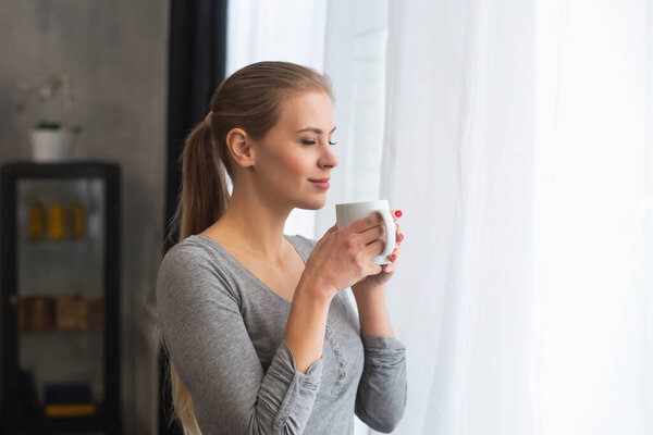 Young teenage girl is standing in front of the window and looking into it. Blond woman at home.