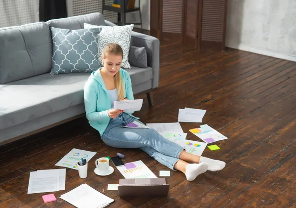 Молодая женщина работает с документами, используя ноутбук, сидя дома на полу. Студентка, предприниматель или девушка-фрилансер, работающая или обучающаяся дистанционно через интернет. — стоковое фото