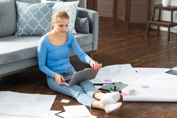 Mujer joven trabaja con documentos utilizando un ordenador portátil mientras está sentado en el suelo en casa. Estudiante, empresaria o freelancer que trabaja o estudia de forma remota a través de Internet. — Foto de Stock