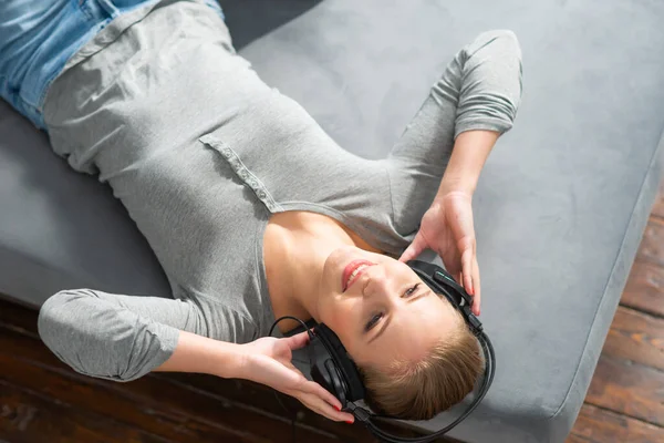 Jovem menina bonita encontra-se em um sofá em fones de ouvido e ouve música. Mulher loira feliz está descansando em casa. — Fotografia de Stock