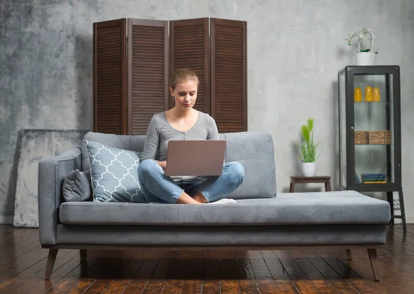 Die junge Frau arbeitet mit einem Laptop, der auf der Couch liegt. Studentin, Unternehmerin oder Freiberuflerin, die aus der Ferne über das Internet arbeitet oder studiert. — Stockfoto