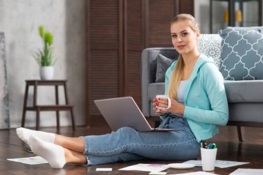 Genç bir kadın evde yerde otururken dizüstü bilgisayar kullanarak belgelerle çalışıyor. Öğrenci, girişimci veya serbest çalışan kız İnternet üzerinden çalışıyor veya uzaktan öğrenim görüyor.