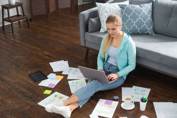 Молода жінка працює з документами, використовуючи ноутбук, сидячи на підлозі вдома. Студент, підприємець або дівчина-фрілансер, яка працює або навчається дистанційно через Інтернет . — стокове фото