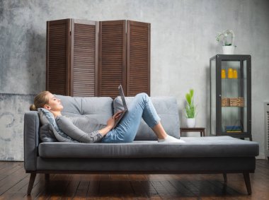 Genç bir kadın kanepede dizüstü bilgisayar kullanarak çalışıyor. Öğrenci, girişimci veya serbest çalışan kız İnternet üzerinden çalışıyor veya uzaktan öğrenim görüyor.