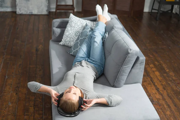 Young beautiful girl lies on a sofa in headphones and listens to music. Happy blond woman is resting at home. — Stock Photo, Image