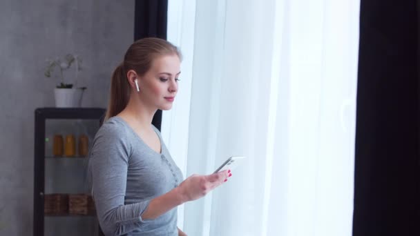 Jong tienermeisje staat met een smartphone voor het raam en kijkt ernaar. Vrouw thuis in de woonkamer. — Stockvideo