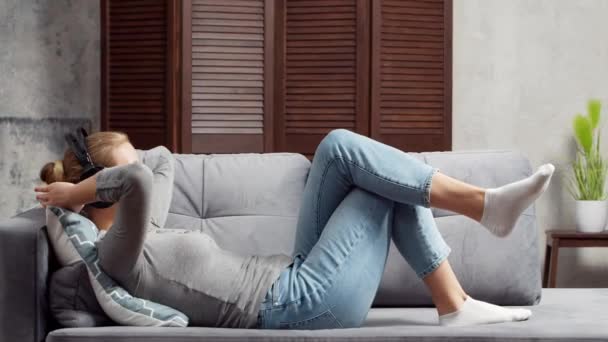 Joven chica hermosa se encuentra en un sofá en los auriculares y escucha música. Mujer feliz está descansando en casa. — Vídeos de Stock