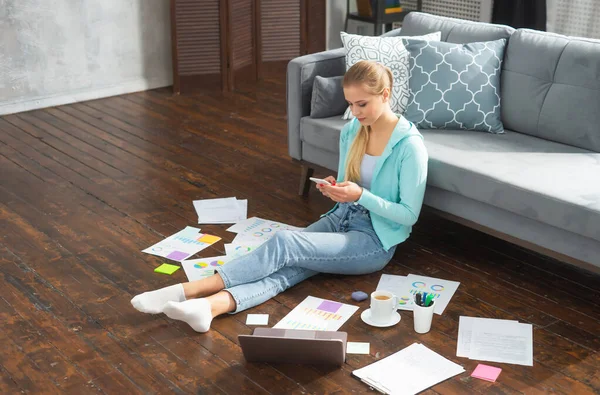Mujer joven trabaja con documentos utilizando teléfono inteligente y portátil mientras está sentado en el suelo en casa. Estudiante, empresaria o freelancer que trabaja o estudia de forma remota a través de Internet. —  Fotos de Stock