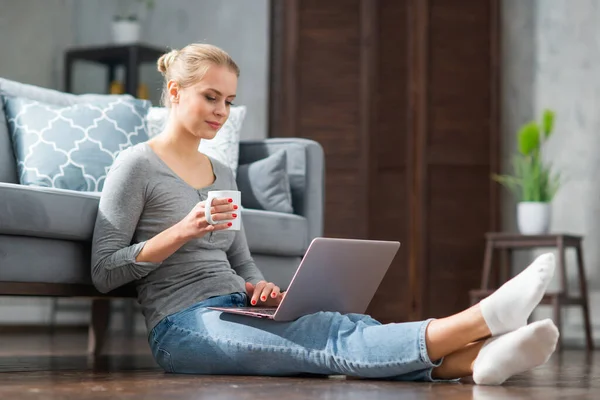Молодая женщина работает с ноутбуком, сидя дома на полу. Студентка, предприниматель или девушка-фрилансер, работающая или обучающаяся дистанционно через интернет. — стоковое фото