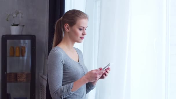 Une jeune adolescente se tient debout avec un smartphone devant la fenêtre et regarde dedans. Femme à la maison dans le salon. — Video