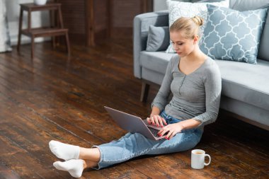 Genç bir kadın evde yerde otururken dizüstü bilgisayar kullanıyor. Öğrenci, girişimci veya serbest çalışan kız İnternet üzerinden çalışıyor veya uzaktan öğrenim görüyor.