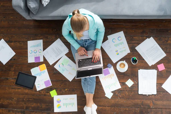 Giovane donna lavora con i documenti utilizzando un computer portatile mentre seduto sul pavimento a casa. Studente, imprenditrice o freelance che lavora o studia da remoto via internet. — Foto Stock