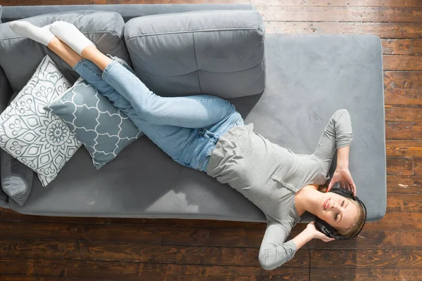 Ung vacker flicka ligger på en soffa i hörlurar och lyssnar på musik. Glad blond kvinna vilar hemma. — Stockfoto