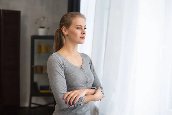 Ein junges Teenager-Mädchen steht vor dem Fenster und schaut hinein. Blonde Frau zu Hause. — Stockfoto