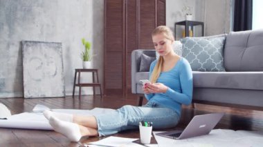 Kadın, evde yerde otururken akıllı telefon ve dizüstü bilgisayar kullanarak belgelerle çalışıyor. Öğrenci, girişimci ya da serbest çalışan ya da uzaktan eğitim gören bir kız. Mesafe öğrenme ve iş kavramı.
