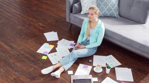 Mujer joven trabaja con documentos utilizando un ordenador portátil mientras está sentado en el suelo en casa. Estudiante, empresaria o freelancer que trabaja o estudia a distancia. Concepto de aprendizaje a distancia y empleo. — Vídeos de Stock