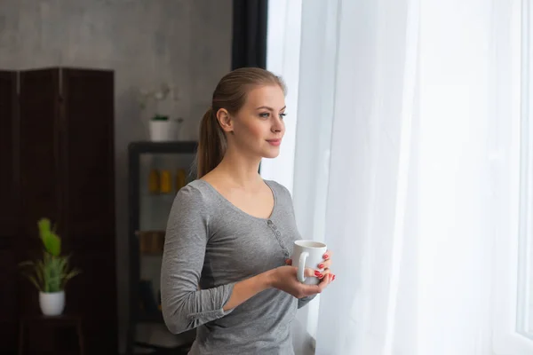 Het jonge tienermeisje staat voor het raam en kijkt ernaar. Blond vrouw thuis. — Stockfoto