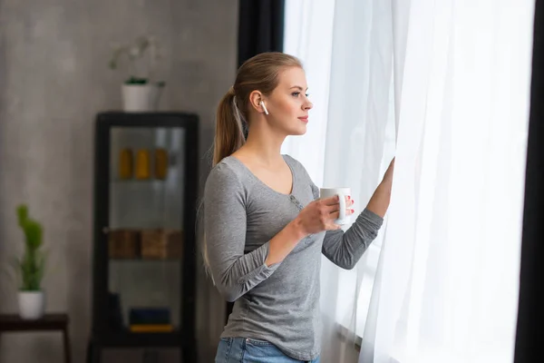 Unga tonårstjej står framför fönstret och tittar in i det. Blond kvinna hemma. — Stockfoto