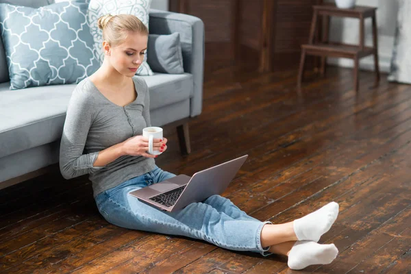 年轻妇女坐在家里的地板上，一边用笔记本电脑工作。通过因特网从事远程工作或学习的学生、企业家或自由职业者. — 图库照片